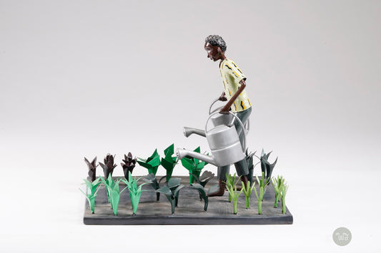 A gardener in a greenish shirt walks barefoot through vegetable beds with two silver watering cans to water the plants