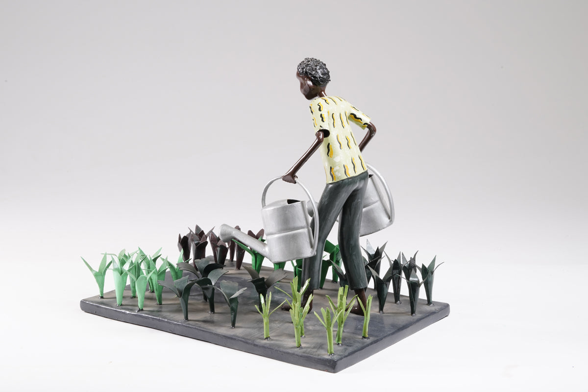 A gardener in a greenish shirt walks barefoot through vegetable beds with two silver watering cans to water the plants