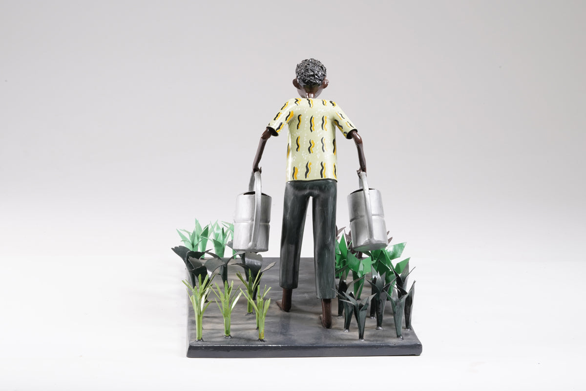 A gardener in a greenish shirt walks barefoot through vegetable beds with two silver watering cans to water the plants