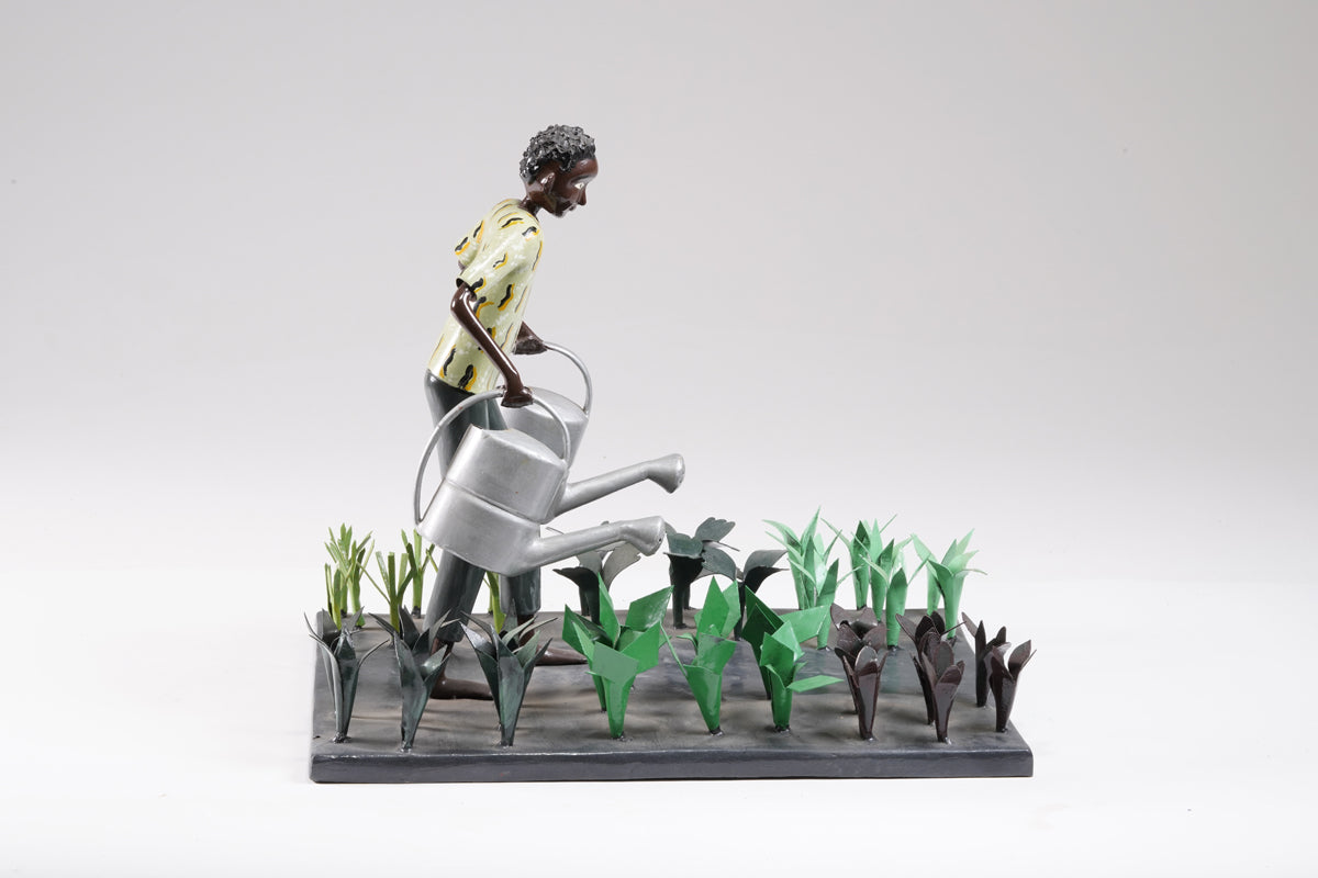 A gardener in a greenish shirt walks barefoot through vegetable beds with two silver watering cans to water the plants