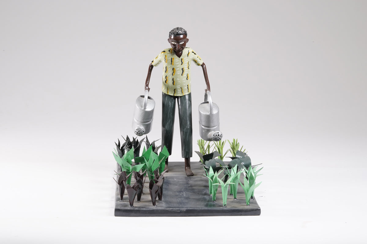 A gardener in a greenish shirt walks barefoot through vegetable beds with two silver watering cans to water the plants