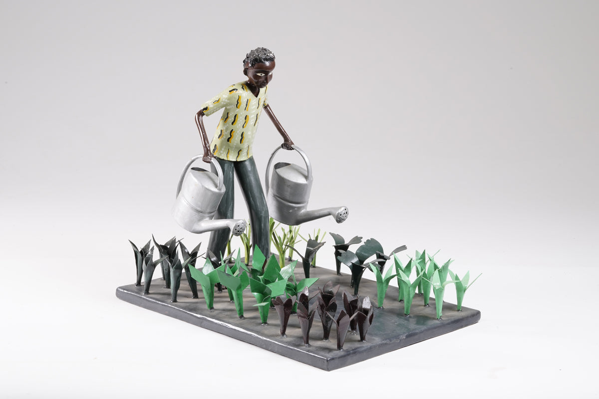 A gardener in a greenish shirt walks barefoot through vegetable beds with two silver watering cans to water the plants