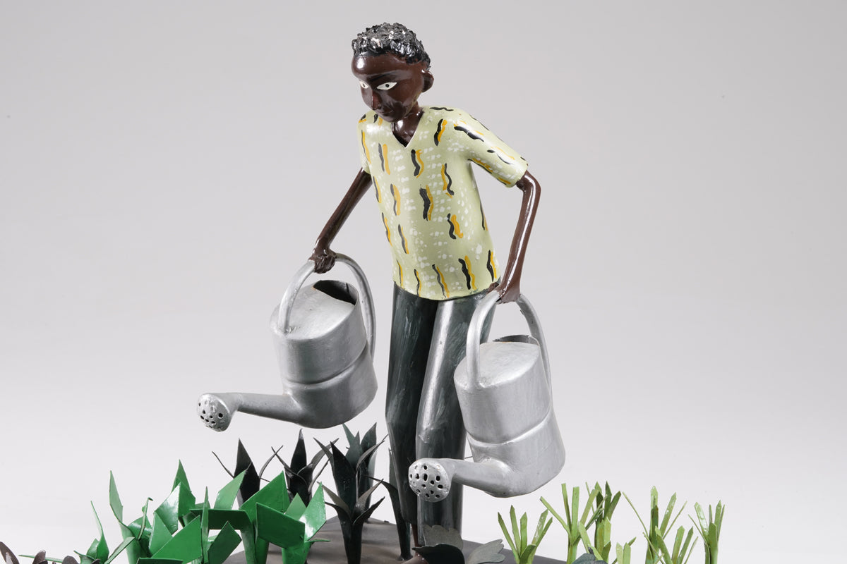 A gardener in a greenish shirt walks barefoot through vegetable beds with two silver watering cans to water the plants