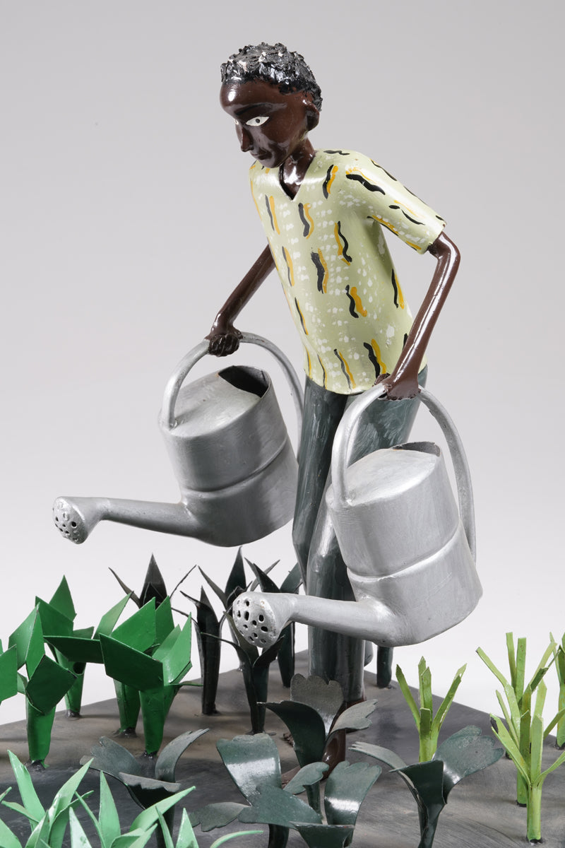 A gardener in a greenish shirt walks barefoot through vegetable beds with two silver watering cans to water the plants