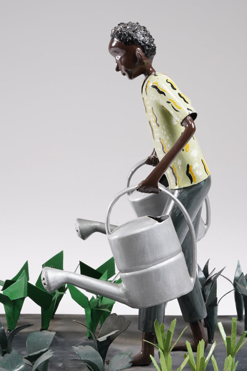A gardener in a greenish shirt walks barefoot through vegetable beds with two silver watering cans to water the plants