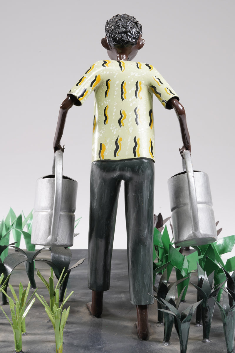 A gardener in a greenish shirt walks barefoot through vegetable beds with two silver watering cans to water the plants