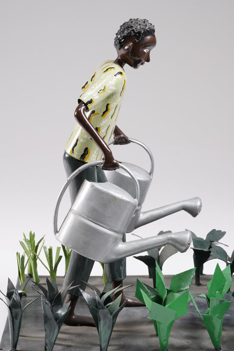 A gardener in a greenish shirt walks barefoot through vegetable beds with two silver watering cans to water the plants