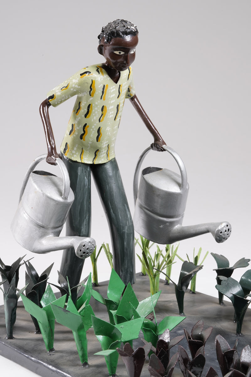 A gardener in a greenish shirt walks barefoot through vegetable beds with two silver watering cans to water the plants