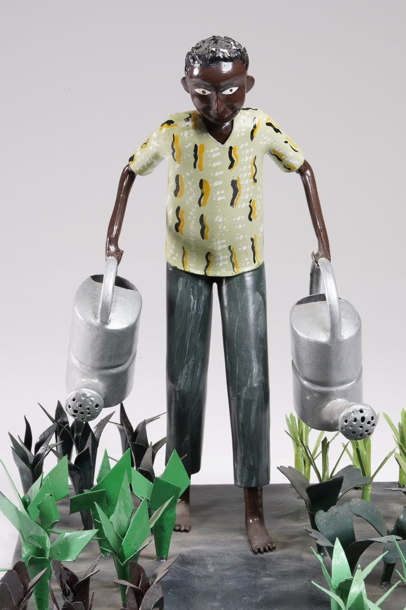A gardener in a greenish shirt walks barefoot through vegetable beds with two silver watering cans to water the plants