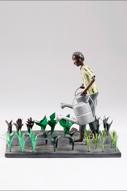 A gardener in a greenish shirt walks barefoot through vegetable beds with two silver watering cans to water the plants
